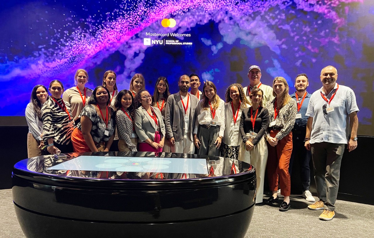 A group of AAS in Hospitality students stand in front of a digital display that says Mastercard Welcomes NYU School of Professional Studies for an Applied Learning event